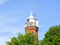 Widok na ceglaną latarnię morską w Ustce z tarasem widokowym, na którym stoją ludzie. W tle błękitne niebo i zielone drzewa.