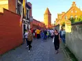 Tłum ludzi spaceruje brukowaną uliczką w Lęborku, obok zabytkowych murów i wieży. Może szukają informacji o lębork zamek cennik?
