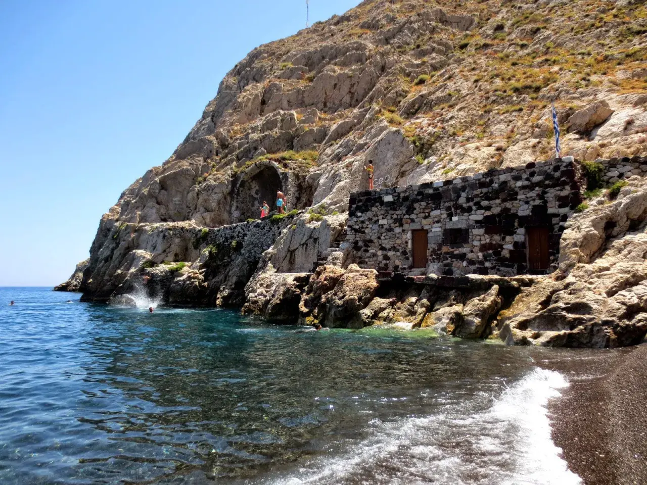Skaliste klify i kamienna budowla nad turkusową wodą, gdzie ludzie skaczą do morza. To urokliwa, czerwona plaża Santorini.