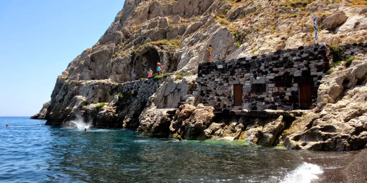 Skaliste klify i kamienna budowla nad turkusową wodą, gdzie ludzie skaczą do morza. To urokliwa, czerwona plaża Santorini.
