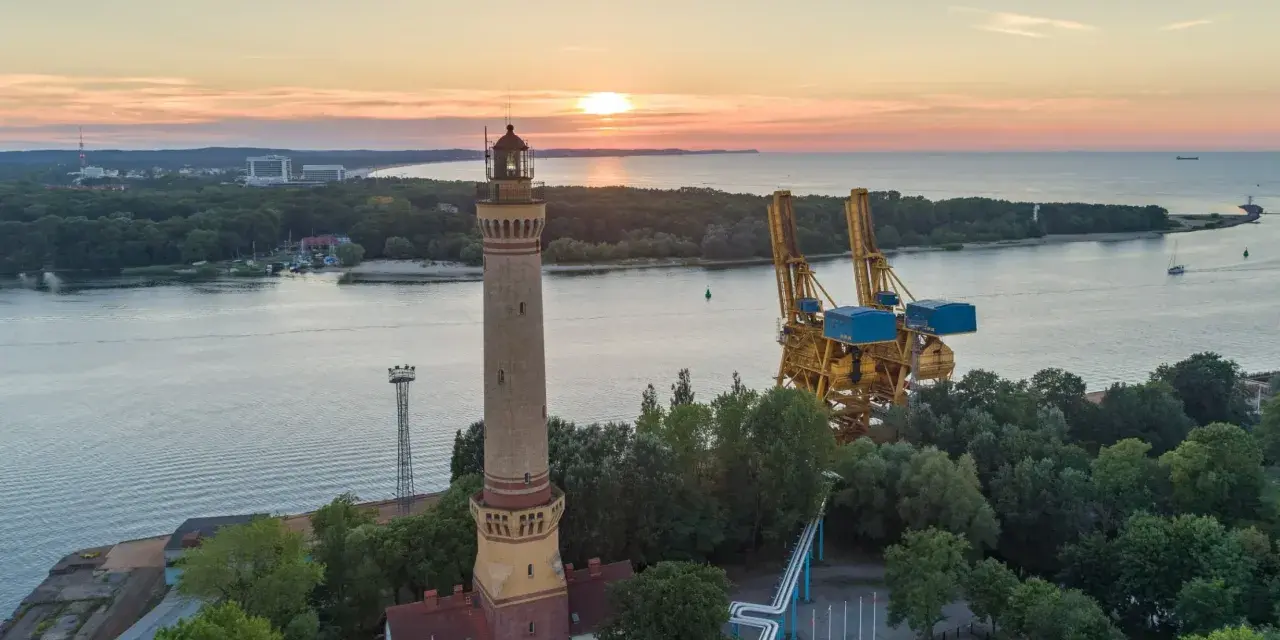 Zachód słońca nad morzem, obok najwyższej latarni morskiej w Polsce, widać też portowe dźwigi.