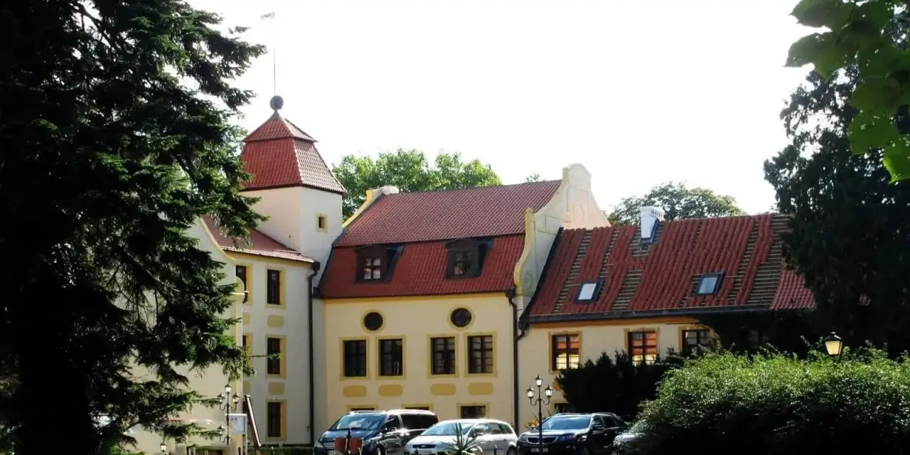Zamek z czerwoną wieżą i dachami, otoczony zielenią. Samochody zaparkowane na trawniku. Idealne miejsce na krokowa atrakcje.
