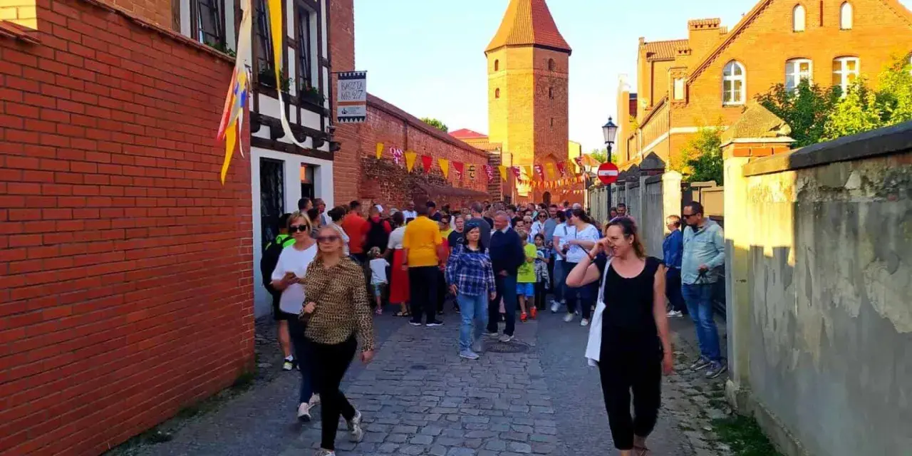 Tłum ludzi spaceruje brukowaną uliczką w Lęborku, obok zabytkowych murów i wieży. Może szukają informacji o lębork zamek cennik?