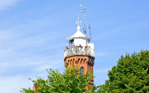 Widok na ceglaną latarnię morską w Ustce z tarasem widokowym, na którym stoją ludzie. W tle błękitne niebo i zielone drzewa.