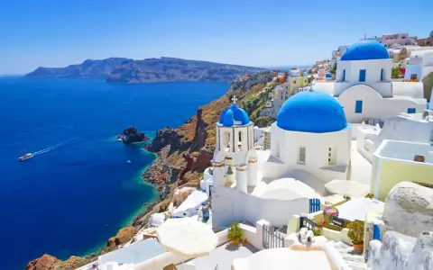 Santorini wycieczki: białe domy z niebieskimi kopułami na klifie nad błękitnym morzem. Statek płynie w oddali.