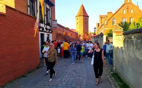 Tłum ludzi spaceruje brukowaną uliczką w Lęborku, obok zabytkowych murów i wieży. Może szukają informacji o lębork zamek cennik?