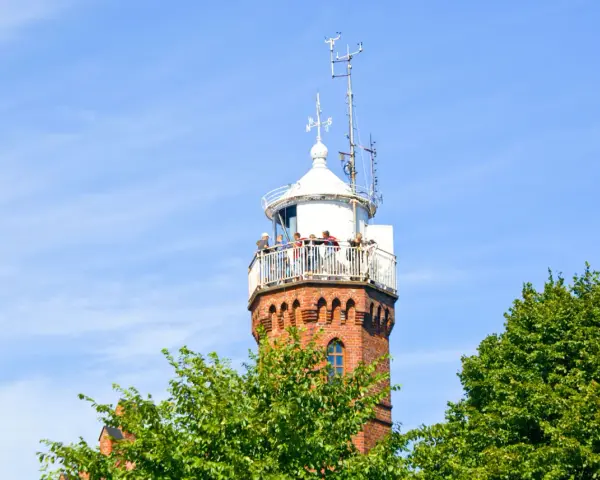 Widok na ceglaną latarnię morską w Ustce z tarasem widokowym, na którym stoją ludzie. W tle błękitne niebo i zielone drzewa.