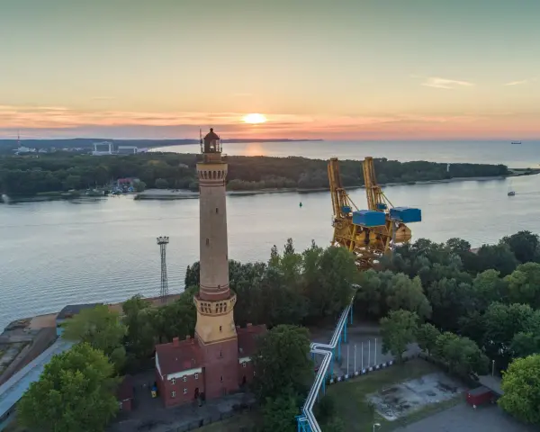 Zachód słońca nad morzem, obok najwyższej latarni morskiej w Polsce, widać też portowe dźwigi.