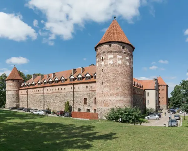 Bytów zamek krzyżacki, masywna budowla z cegły i kamienia z charakterystycznymi wieżami i czerwonymi dachami, otoczona zielenią.