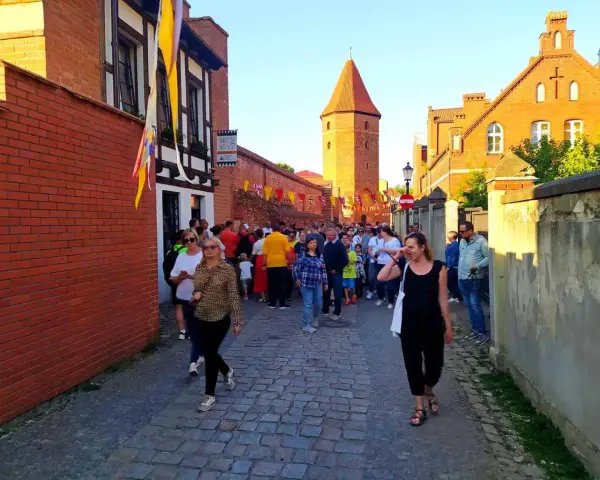 Tłum ludzi spaceruje brukowaną uliczką w Lęborku, obok zabytkowych murów i wieży. Może szukają informacji o lębork zamek cennik?