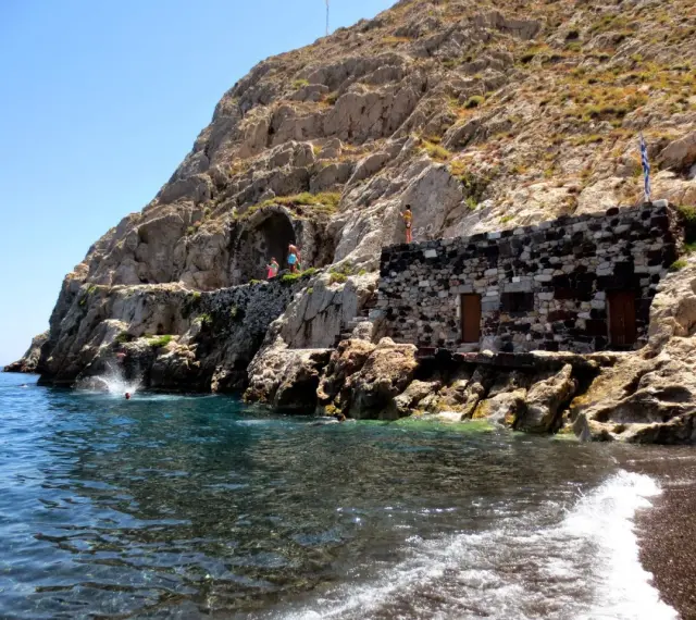 Skaliste klify i kamienna budowla nad turkusową wodą, gdzie ludzie skaczą do morza. To urokliwa, czerwona plaża Santorini.