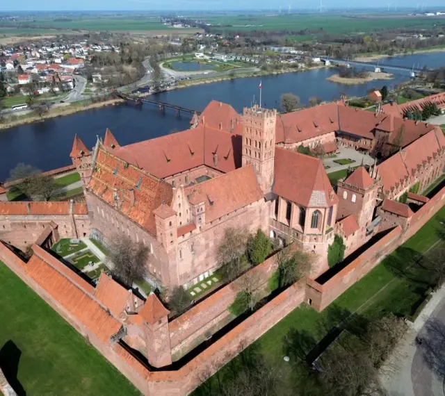 Imponujący zamek w Malborku z lotu ptaka. Rozległa powierzchnia twierdzy z czerwonej cegły otoczona murami obronnymi, z rzeką i miastem w tle.