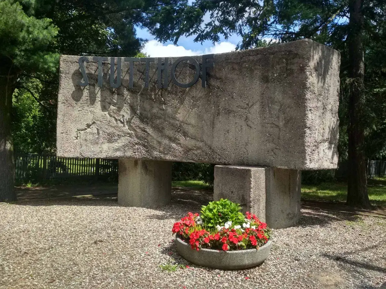 Monumentalny napis STUTTHOF na betonowym obelisku. Ciekawe miejsce pamięci, otoczone zielenią i kwiatami.