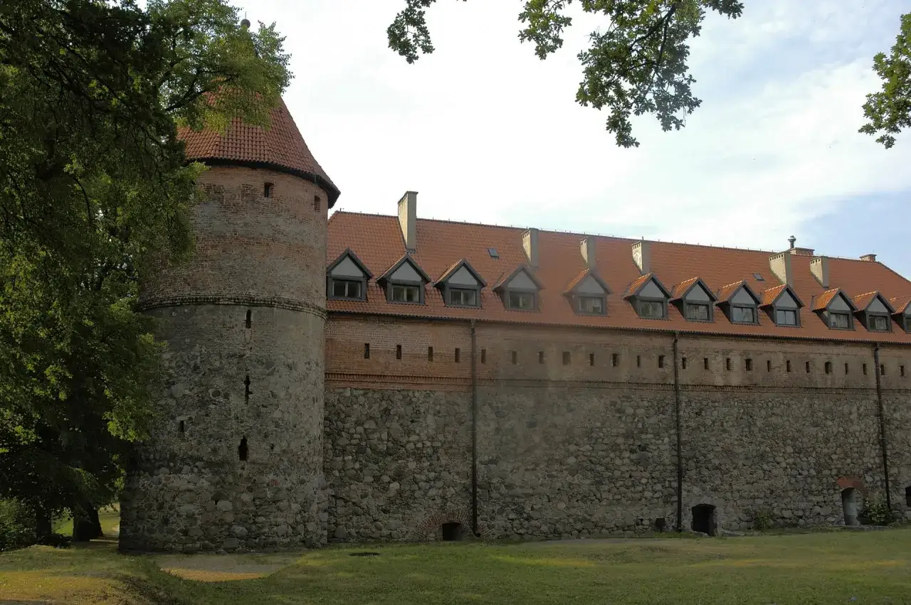 Okazały bytów zamek krzyżacki z okrągłą wieżą i kamiennymi murami, otoczony zielenią.