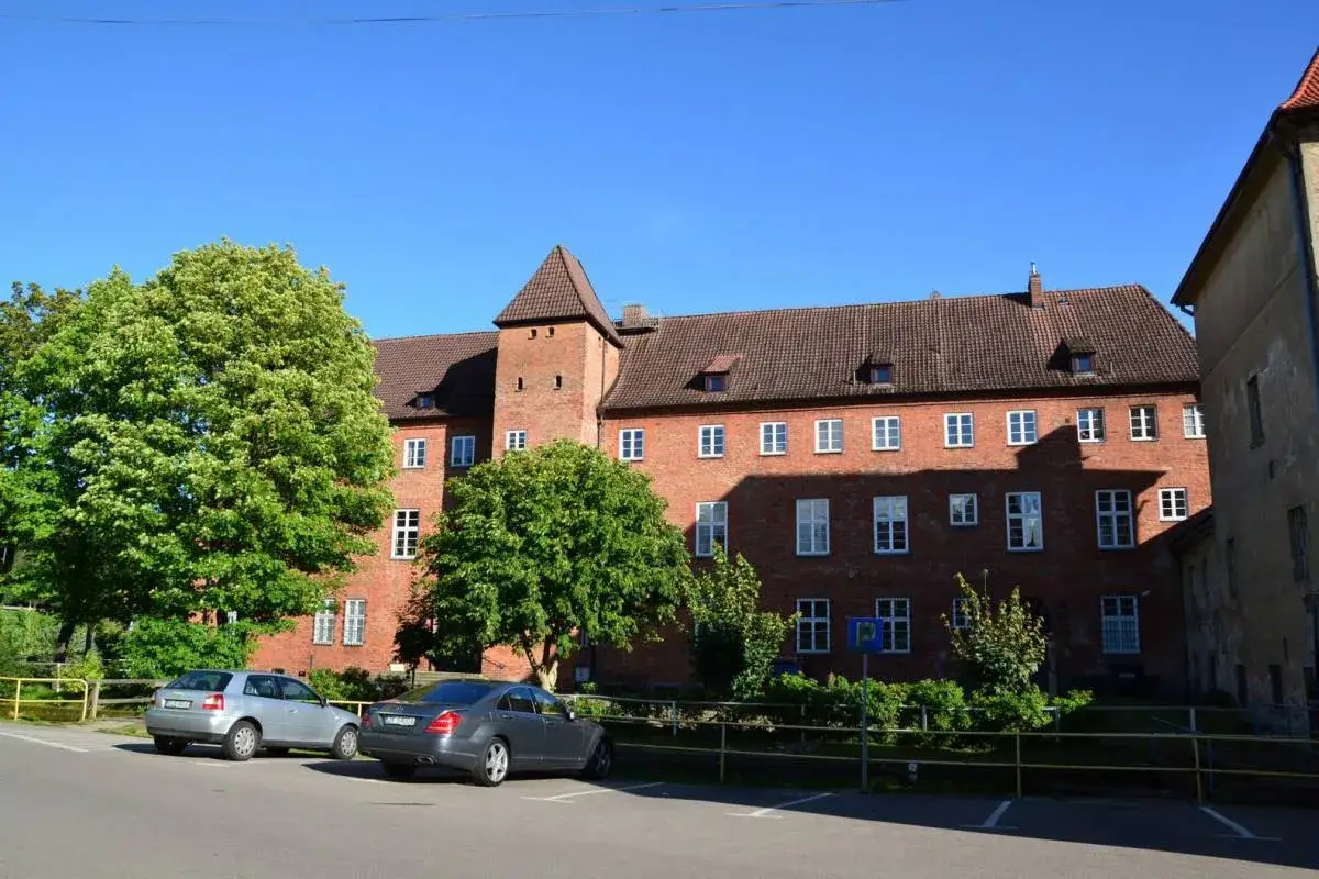 Lębork zamek cennik: ceglany budynek z wieżą, drzewa, dwa samochody na parkingu.