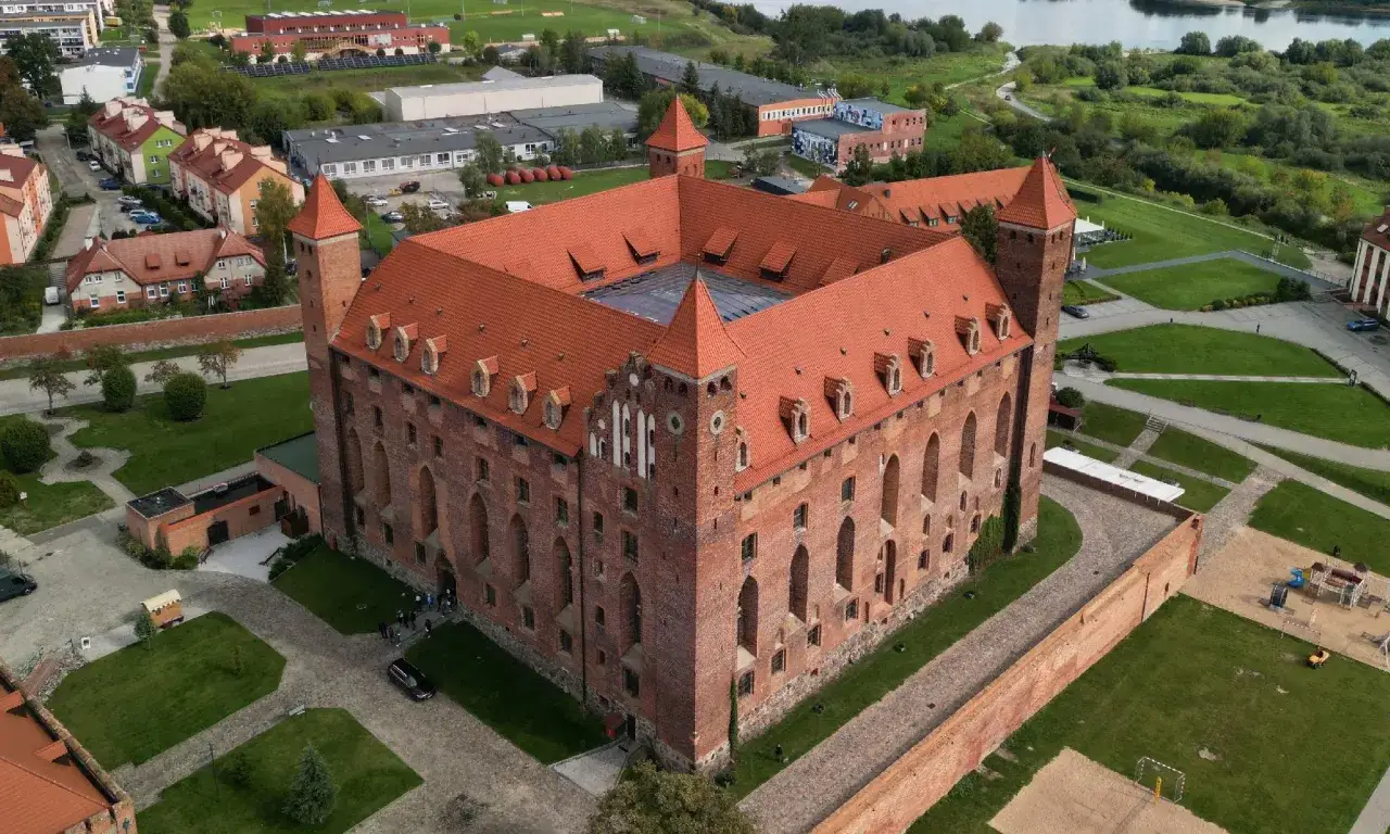 Zamek w Gniewie zaprasza na zwiedzanie. Potężna ceglana twierdza z czerwonymi dachami i wieżami, otoczona zielenią i pobliską zabudową.