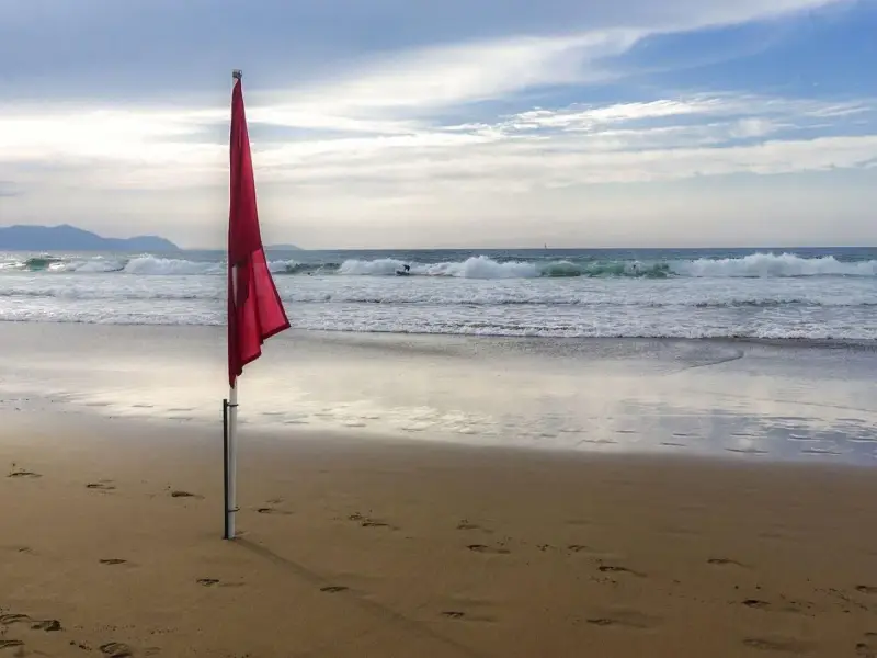 Czerwone flagi na plaży ostrzegają przed niebezpiecznymi falami. Surferzy korzystają z fal, podczas gdy na piasku widać ślady stóp.